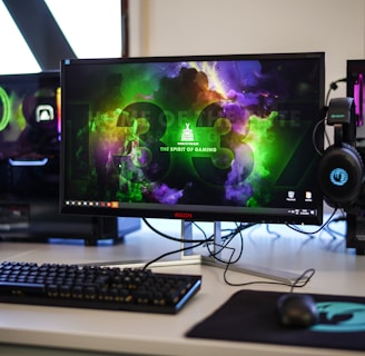 A gaming setup featuring a central monitor with a vibrant display showing 'The Spirit of Gaming'. On either side, there are gaming PCs with illuminated components and transparent cases. Headsets hang from the side of the monitor. The desk also has a black keyboard and mouse, and a branded mousepad.