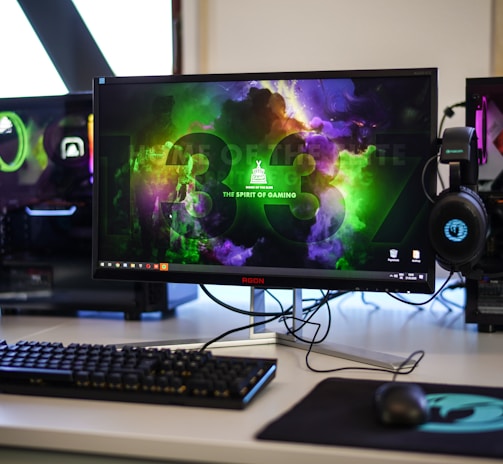 A gaming setup featuring a central monitor with a vibrant display showing 'The Spirit of Gaming'. On either side, there are gaming PCs with illuminated components and transparent cases. Headsets hang from the side of the monitor. The desk also has a black keyboard and mouse, and a branded mousepad.