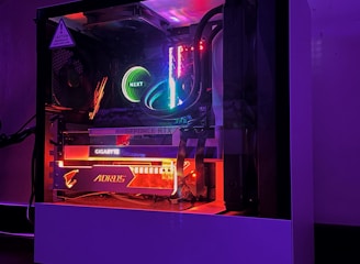 A gaming PC with a transparent side panel reveals internal components illuminated by RGB lighting. Key components visible include a graphics card, cooling system, and motherboard, all surrounded by multicolored LED lights. The case has a minimalistic design, set against a dark background that highlights the vibrant internal lighting.
