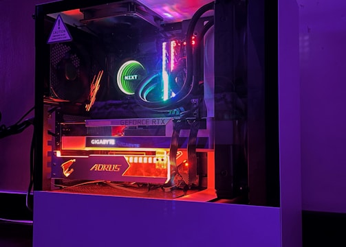 A gaming PC with a transparent side panel reveals internal components illuminated by RGB lighting. Key components visible include a graphics card, cooling system, and motherboard, all surrounded by multicolored LED lights. The case has a minimalistic design, set against a dark background that highlights the vibrant internal lighting.