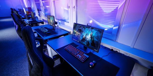 A row of modern gaming setups is arranged neatly along a window-lined wall. Each station includes a high-resolution monitor displaying vivid graphics, a mechanical keyboard with colored lighting, a gaming mouse, and an ergonomic chair. The room is illuminated with ambient blue and purple lights, creating a sleek and futuristic atmosphere.