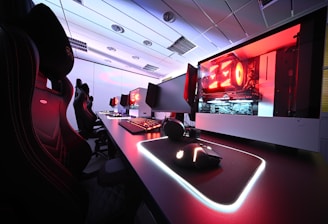 A row of high-end gaming computers on sleek desks, featuring RGB lighting and gaming accessories. The computers are housed in transparent cases with visible components, emitting a red glow. Comfortable gaming chairs with ergonomic design are positioned next to the desks, and the room's ceiling is modern with recessed lighting.