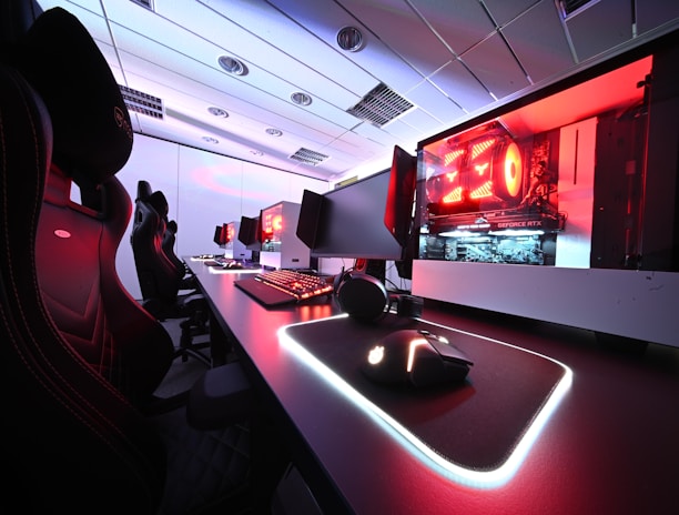 A row of high-end gaming computers on sleek desks, featuring RGB lighting and gaming accessories. The computers are housed in transparent cases with visible components, emitting a red glow. Comfortable gaming chairs with ergonomic design are positioned next to the desks, and the room's ceiling is modern with recessed lighting.