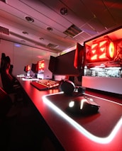 A modern gaming setup featuring several high-end computers with illuminated red interior lighting. The room is equipped with gaming chairs exhibiting a sleek black and red design. Multiple monitors are arranged on a long desk, and peripheral devices like keyboards and mice are uniformly placed, reflecting an ambiance ideal for gaming or competitive play.