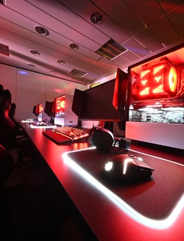 A modern gaming setup featuring several high-end computers with illuminated red interior lighting. The room is equipped with gaming chairs exhibiting a sleek black and red design. Multiple monitors are arranged on a long desk, and peripheral devices like keyboards and mice are uniformly placed, reflecting an ambiance ideal for gaming or competitive play.