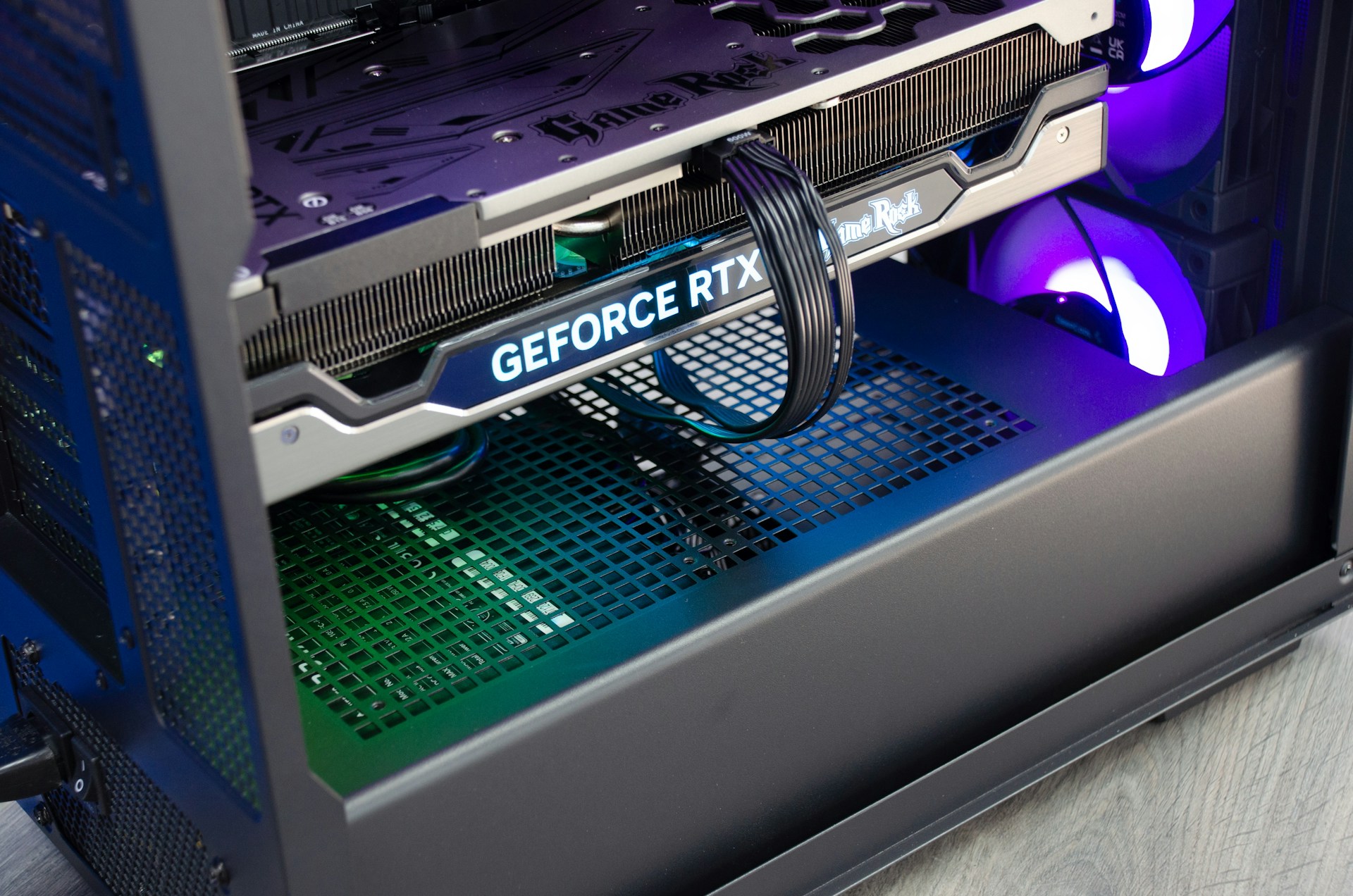 A close-up view of the interior of a computer casing showcasing a high-end graphics card with 'GeForce RTX' branding. The graphics card is secured in its slot, and several power cables are attached. The background reveals cooling fans illuminated with RGB lighting.