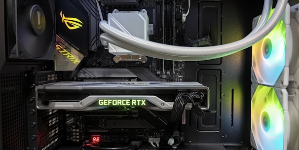 Inside view of a computer case showcasing a sophisticated gaming setup. Visible components include a GeForce RTX graphics card illuminated with green lighting, an ASUS motherboard, cooling fans with LED lighting, and a liquid cooling system with white tubes. Cables are neatly organized, and overall, the hardware suggests a high-performance machine.