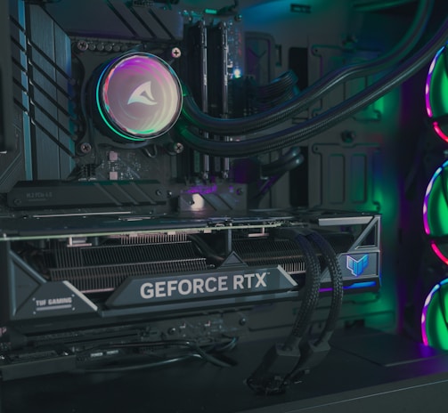 The image depicts the inside of a computer case featuring a high-performance graphics card labeled 'GEFORCE RTX' with RGB lighting. The cooling fans are illuminated with vibrant colors, creating a visually striking look. Various cables and components are neatly arranged, highlighting advanced hardware and a focus on aesthetics.